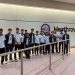 A group of young cricket players in matching MCC uniforms standing together at Heathrow Airport, posing for a team photo near a wall with the Heathrow logo.